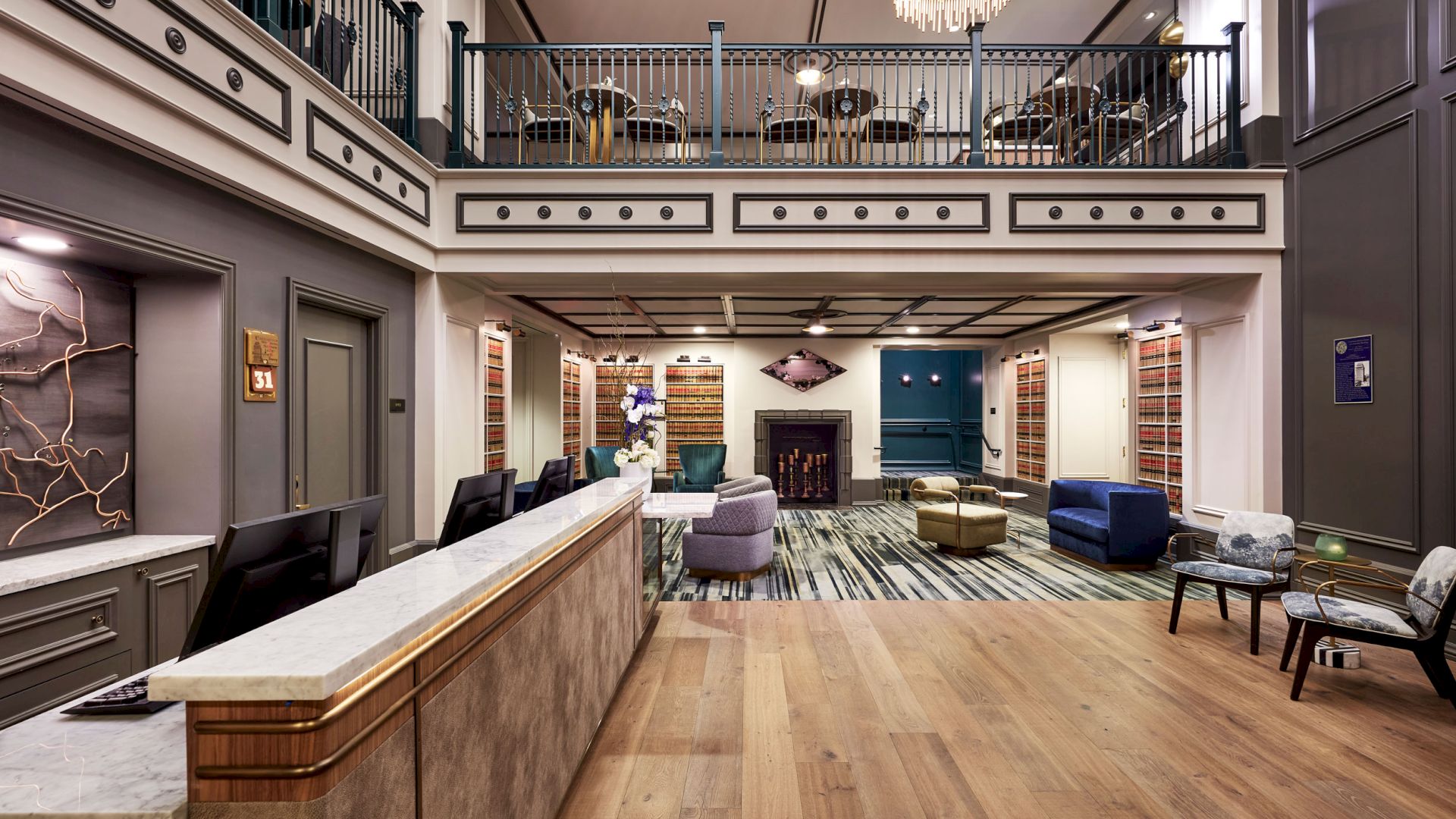 Elegant hotel lounge with marble bar counter, bookshelves, cozy fireplace, upholstered seating, and a grand chandelier over a balcony.