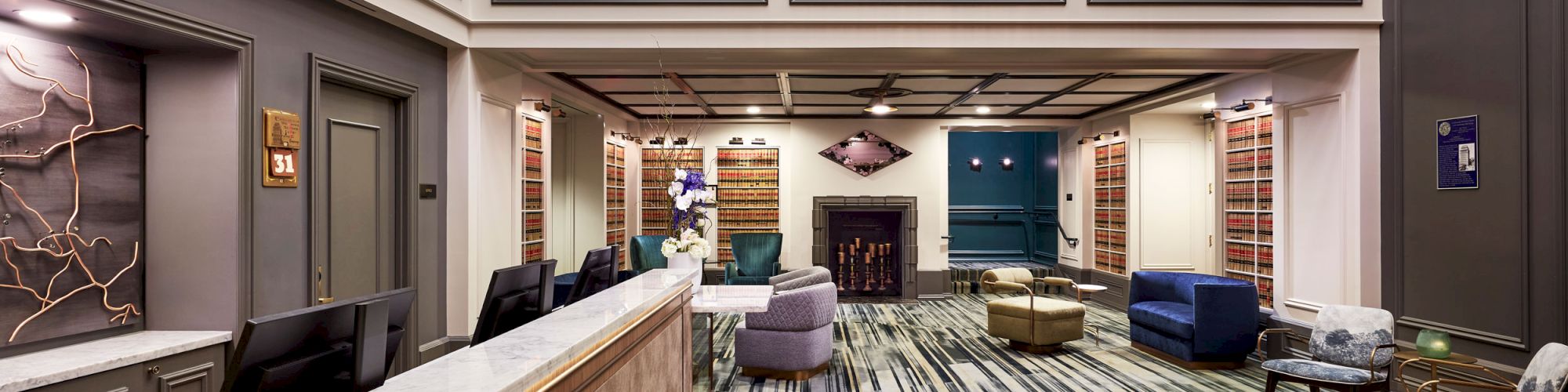 Elegant hotel lounge with marble bar counter, bookshelves, cozy fireplace, upholstered seating, and a grand chandelier over a balcony.