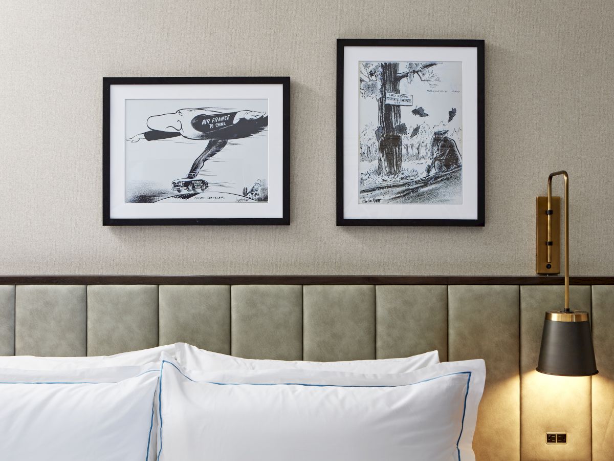 A neatly made bed sits against a tufted headboard beneath two framed black-and-white artworks and a brass wall lamp, glowing warmly.