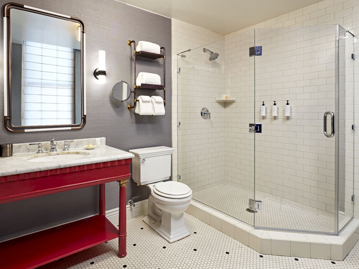 A modern bathroom with a red vanity, mirror, toilet, and a glass shower with tiled walls and towel racks.
