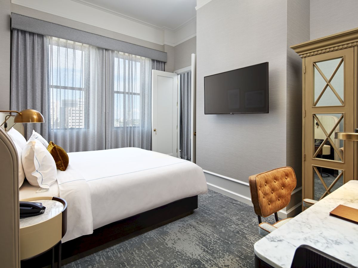 A stylish hotel room with a bed, sheer curtains, a wall-mounted TV, and a polished wooden door with frosted glass panels.