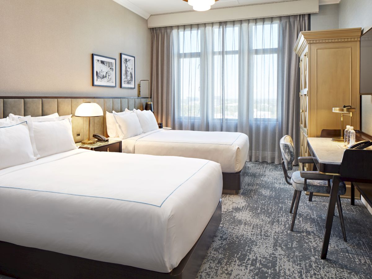 A hotel room with two neatly made beds, bedside lamps, curtains by a window, and a desk with chair.