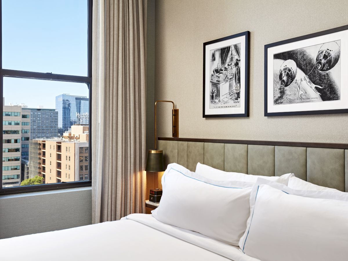 A hotel room corner with a neatly made bed, large window showing a city view, and two framed black-and-white aircraft posters.