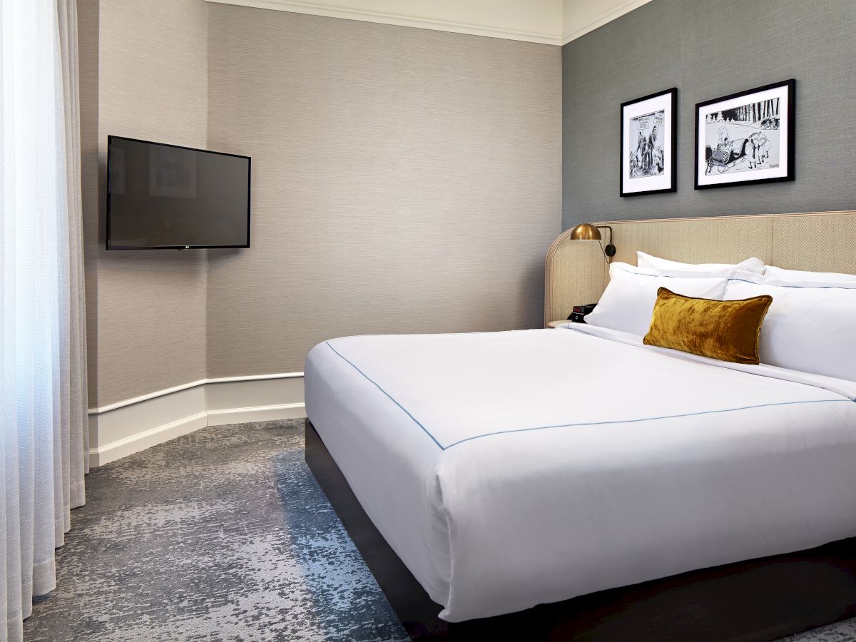 A modern hotel room with a bed, wall-mounted TV, framed art, and a bedside lamp beside curtains.