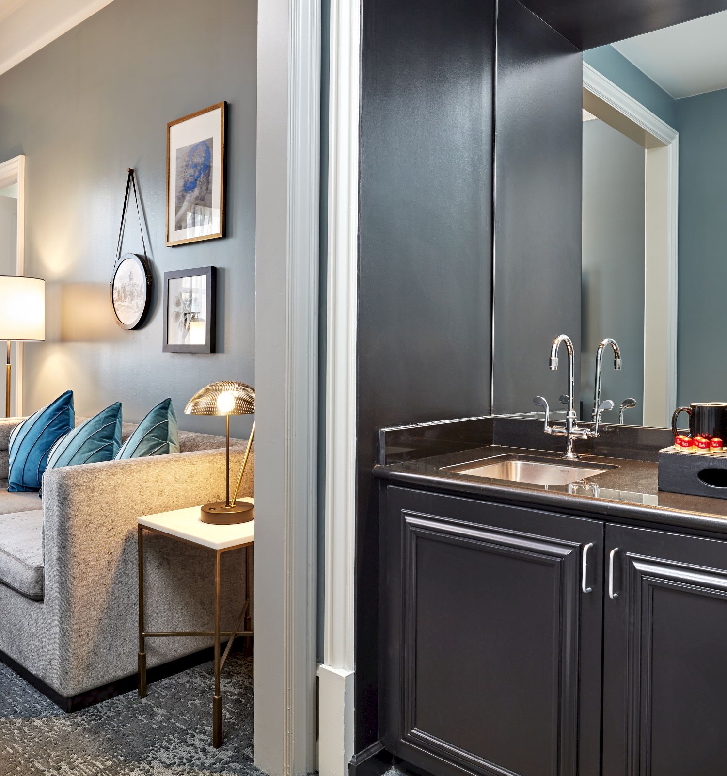 Bright hotel suite view with a gray sofa and lamps, plus a kitchenette counter with sink and coffee maker.