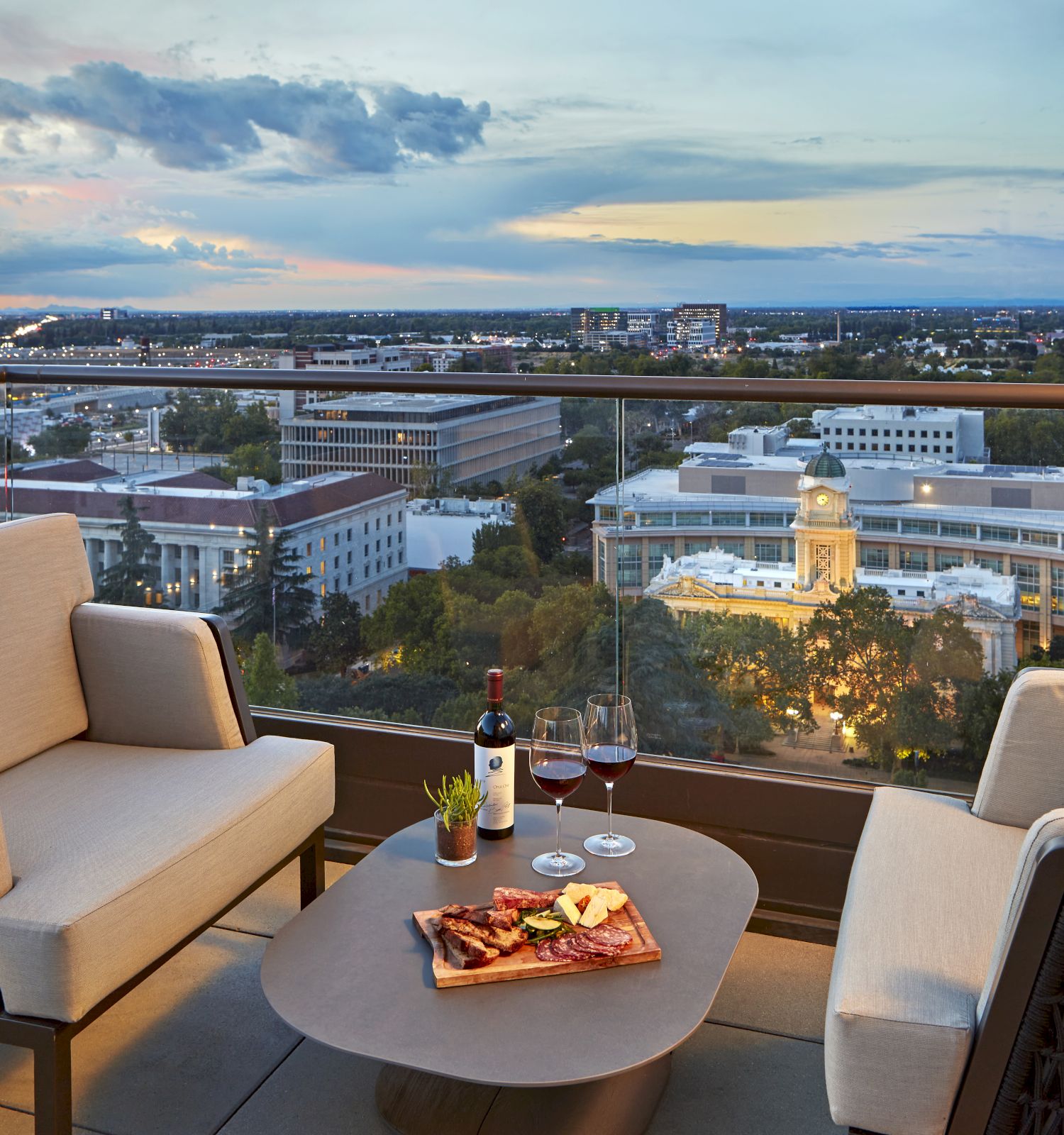 Rooftop balcony dinner with wine and glasses overlooking a city skyline at dusk, with clouds and glowing buildings in the distance.
