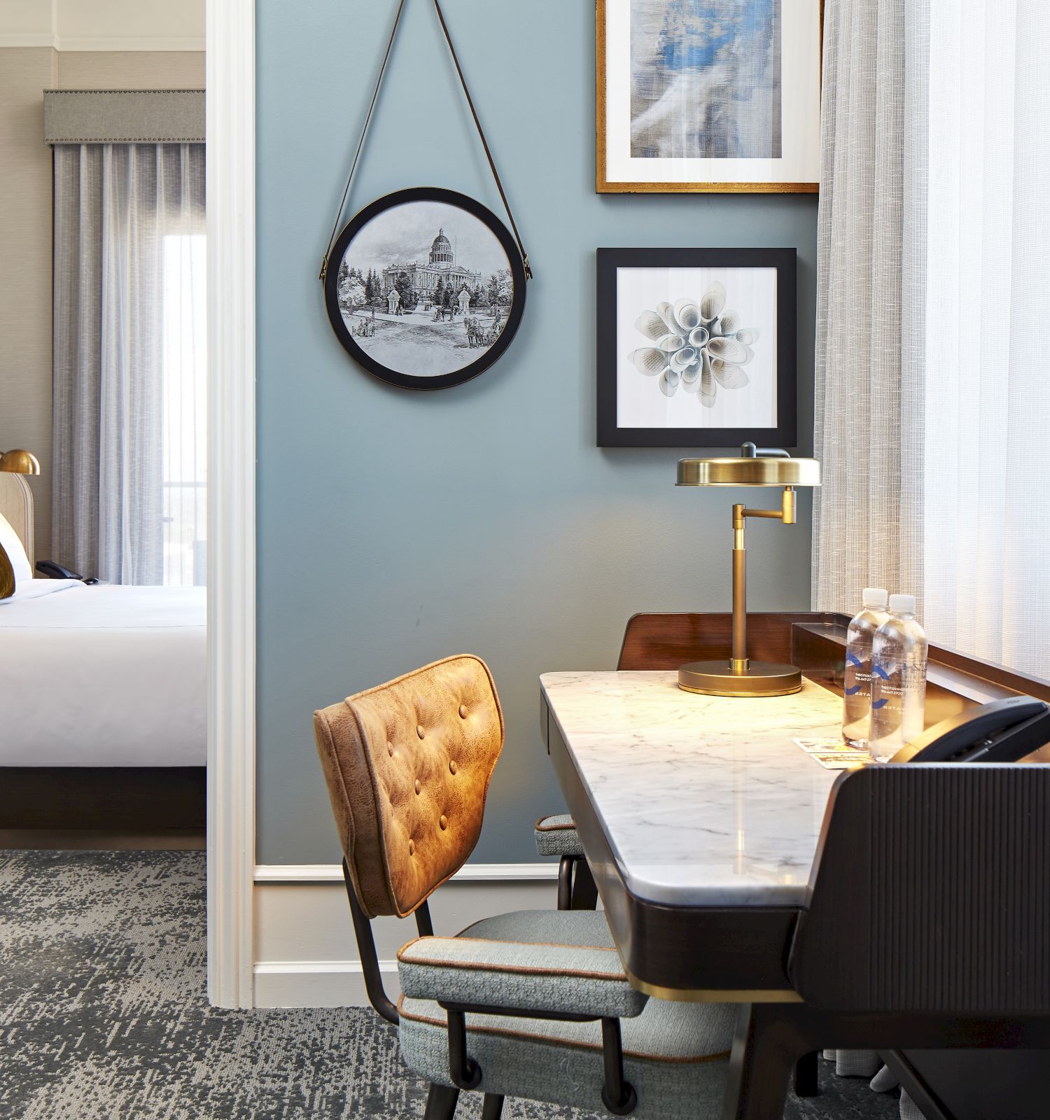 A modern hotel suite shows a bedroom with curtains, plus a desk with lamp and tray, and framed wall art on a teal wall.