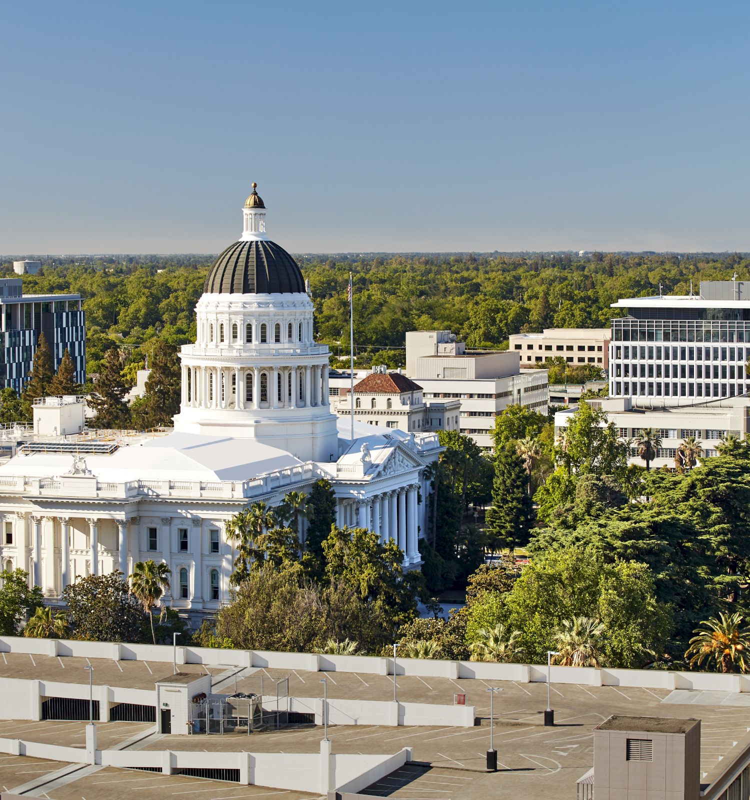 A sunny aerial view of a capital government complex with a large white domed building, surrounding offices, and lush trees, and clear skies.