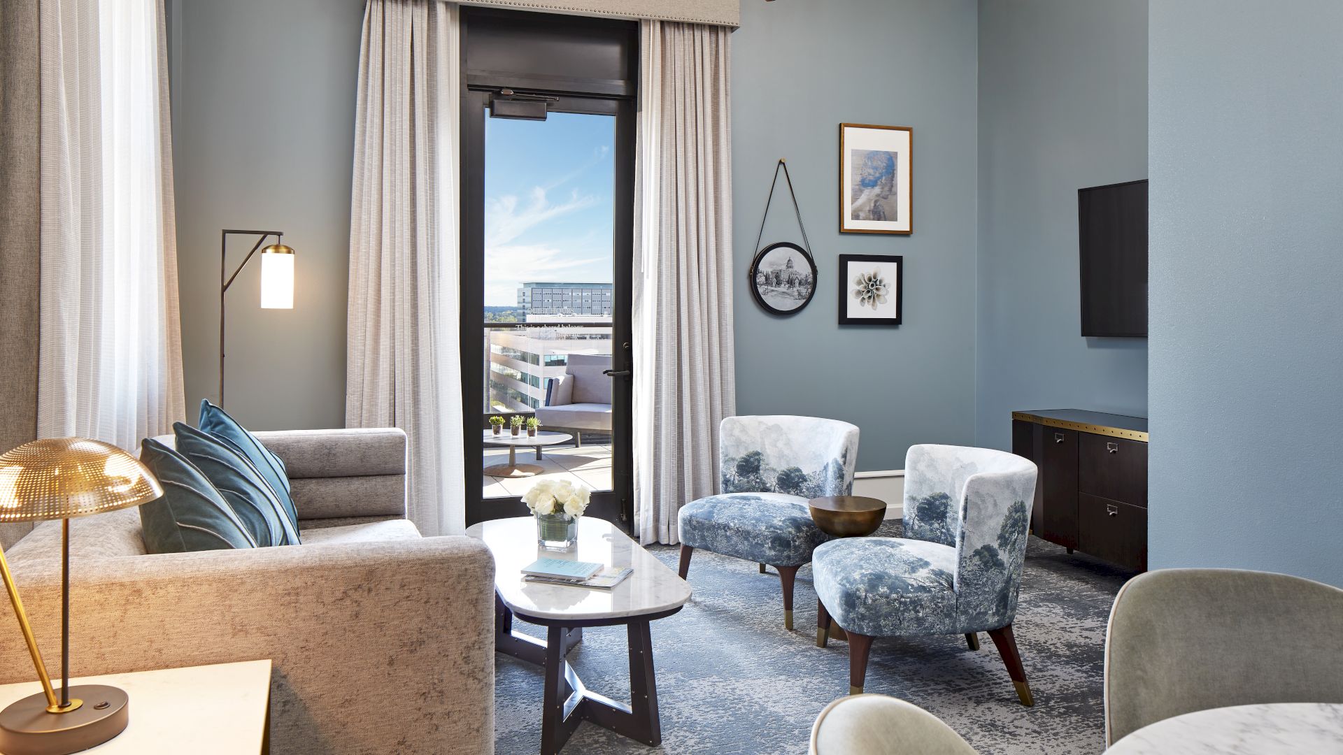 A modern lounge with a sofa, two blue armchairs, coffee table, framed art, TV, and floor-to-ceiling curtains.