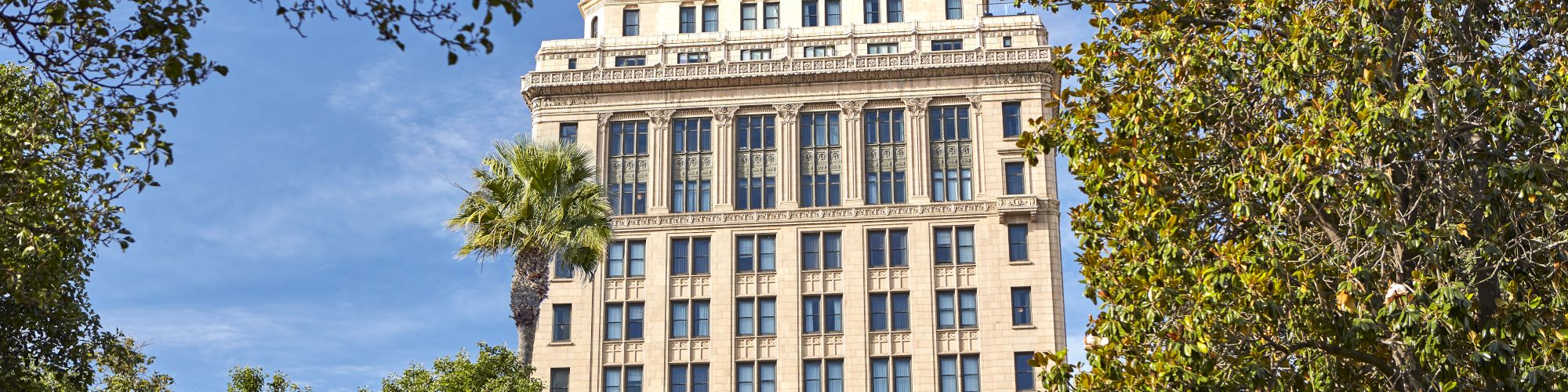 A tall neoclassical office building rises behind leafy trees under a blue sky.