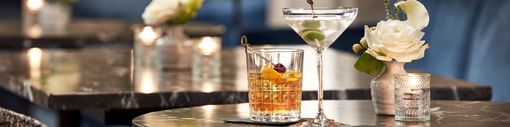 A bar table set with an amber cocktail in a glass, a martini-style drink, and white flowers in a vase, all warmly lit.
