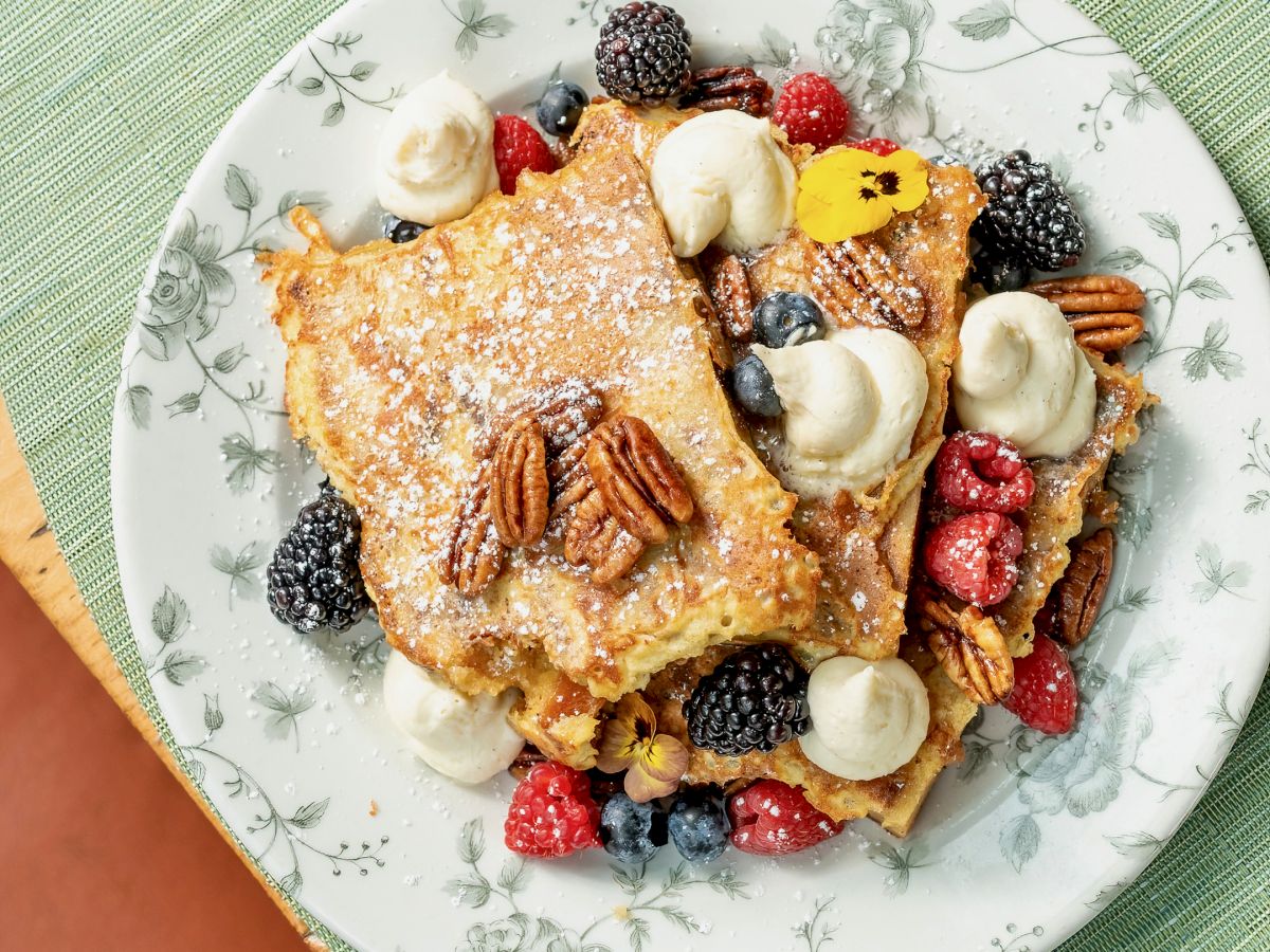 A plate of French toast topped with banana slices, pecans, and mixed berries like raspberries, blueberries, and blackberries.
