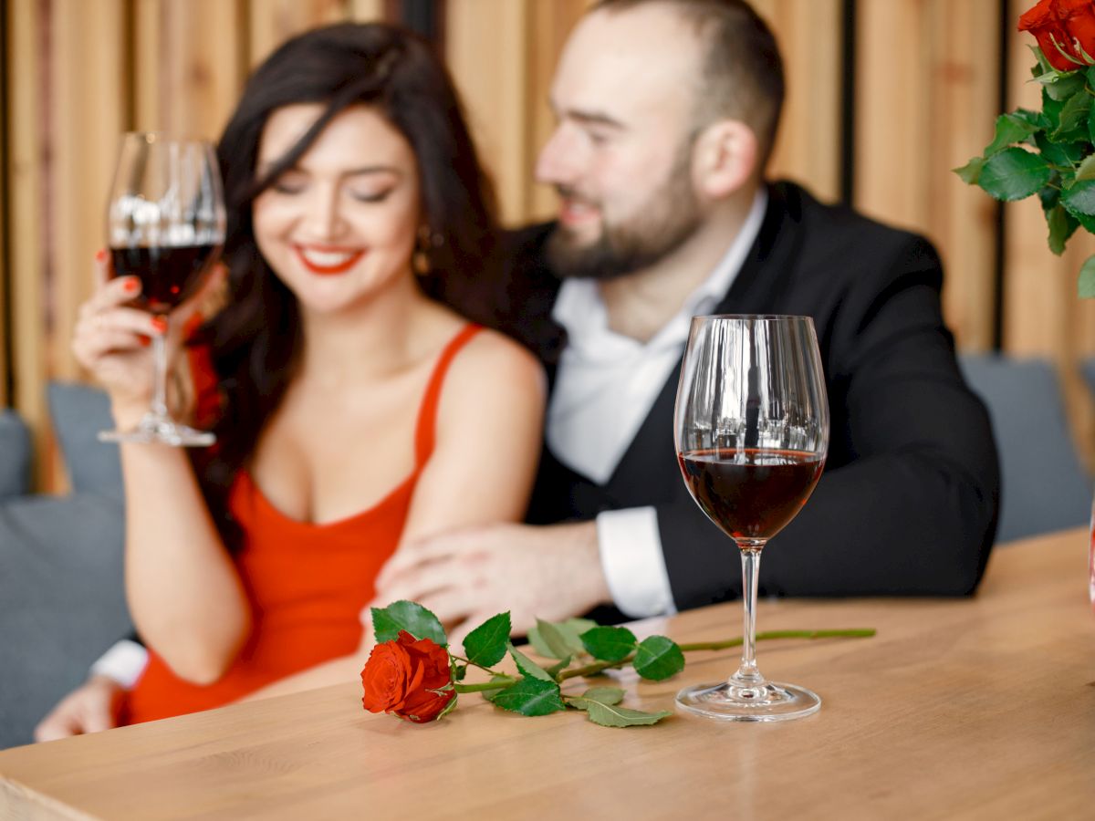 A couple dressed up toast with wine at a table, with a vase of red roses nearby in warm, romantic lighting.