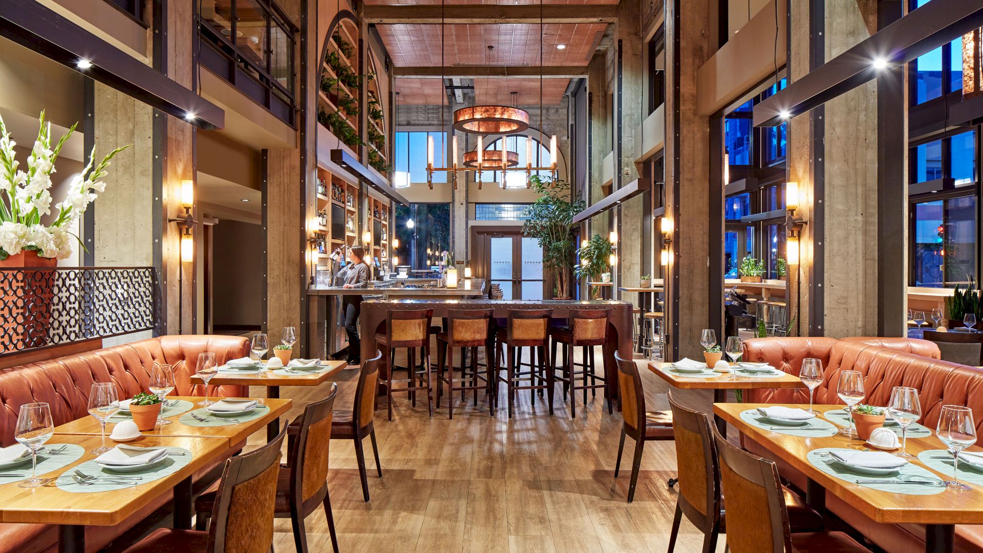 A modern restaurant interior with banquet seating, set tables, warm lights, and a visible bar in the background.
