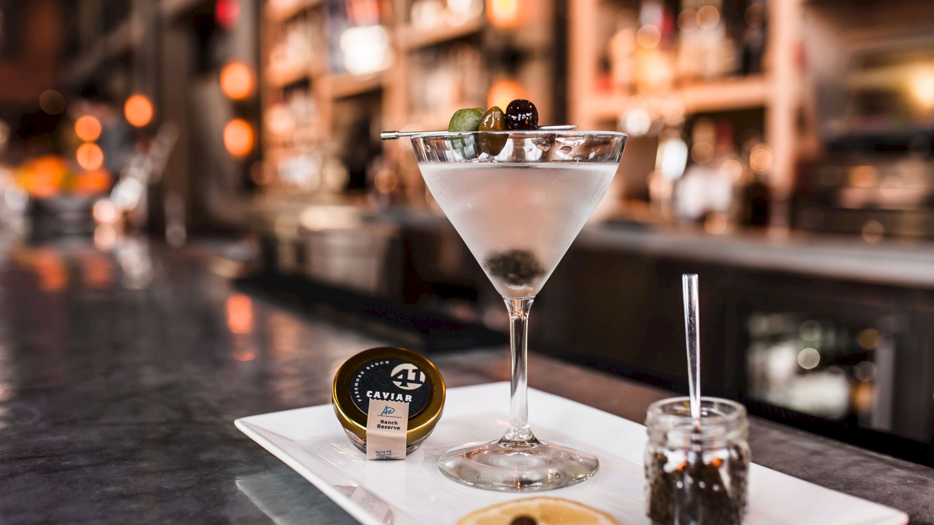 A martini glass with ice and olives, caviar nearby, set on a bar counter with blurred lights behind it.