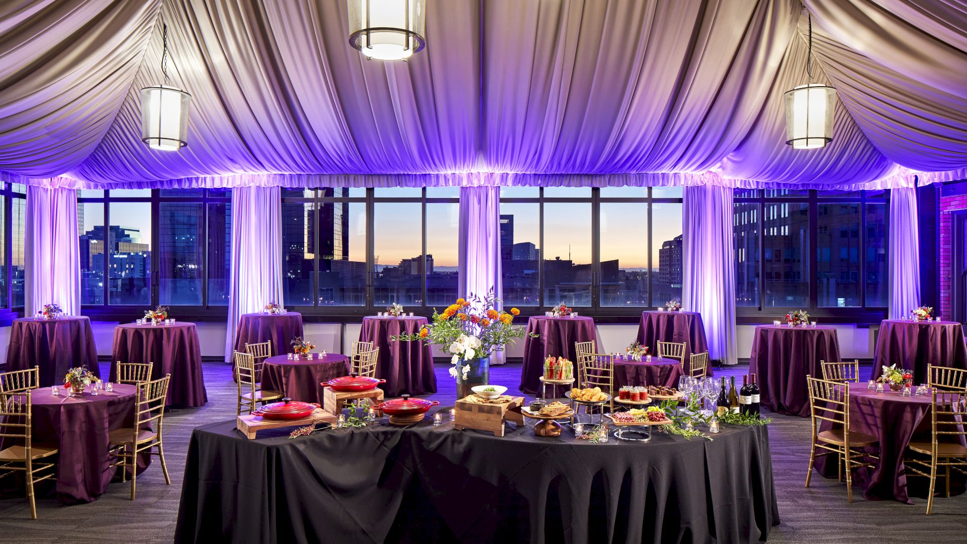 A banquet setup with purple drapes, round tables, chandeliers, and city skyline views at dusk.