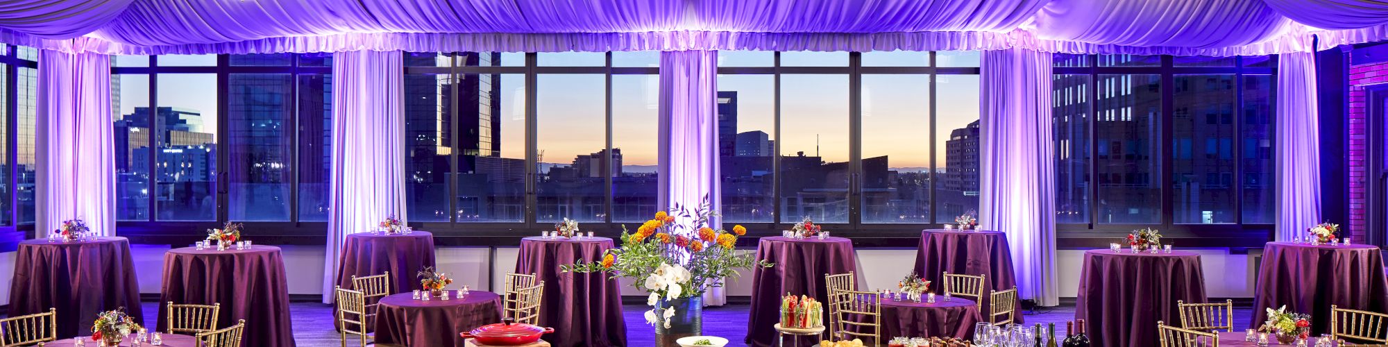 A banquet setup with purple drapes, round tables, chandeliers, and city skyline views at dusk.