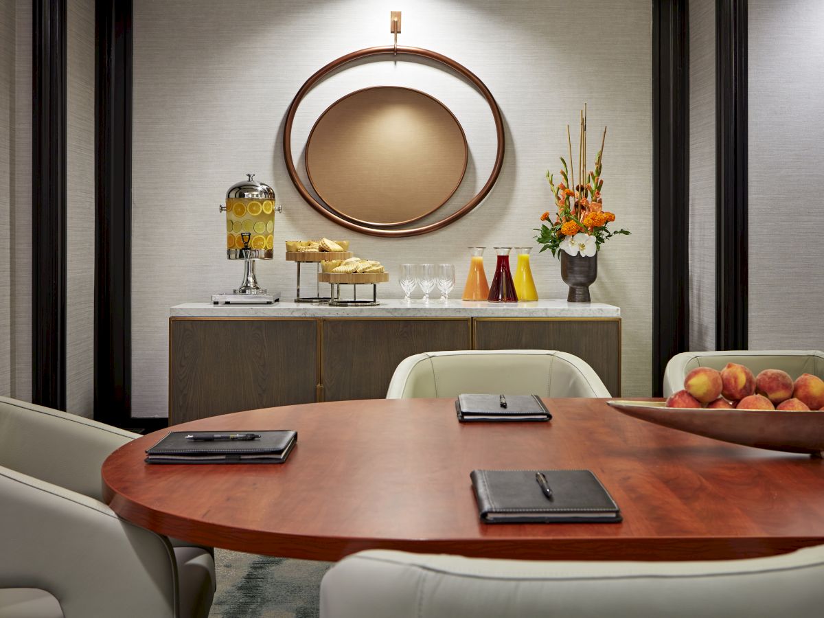 A modern hotel bar/serving counter with a drink dispenser, glasses, colorful bottles, fresh flowers, and a fruit tray in a meeting room.