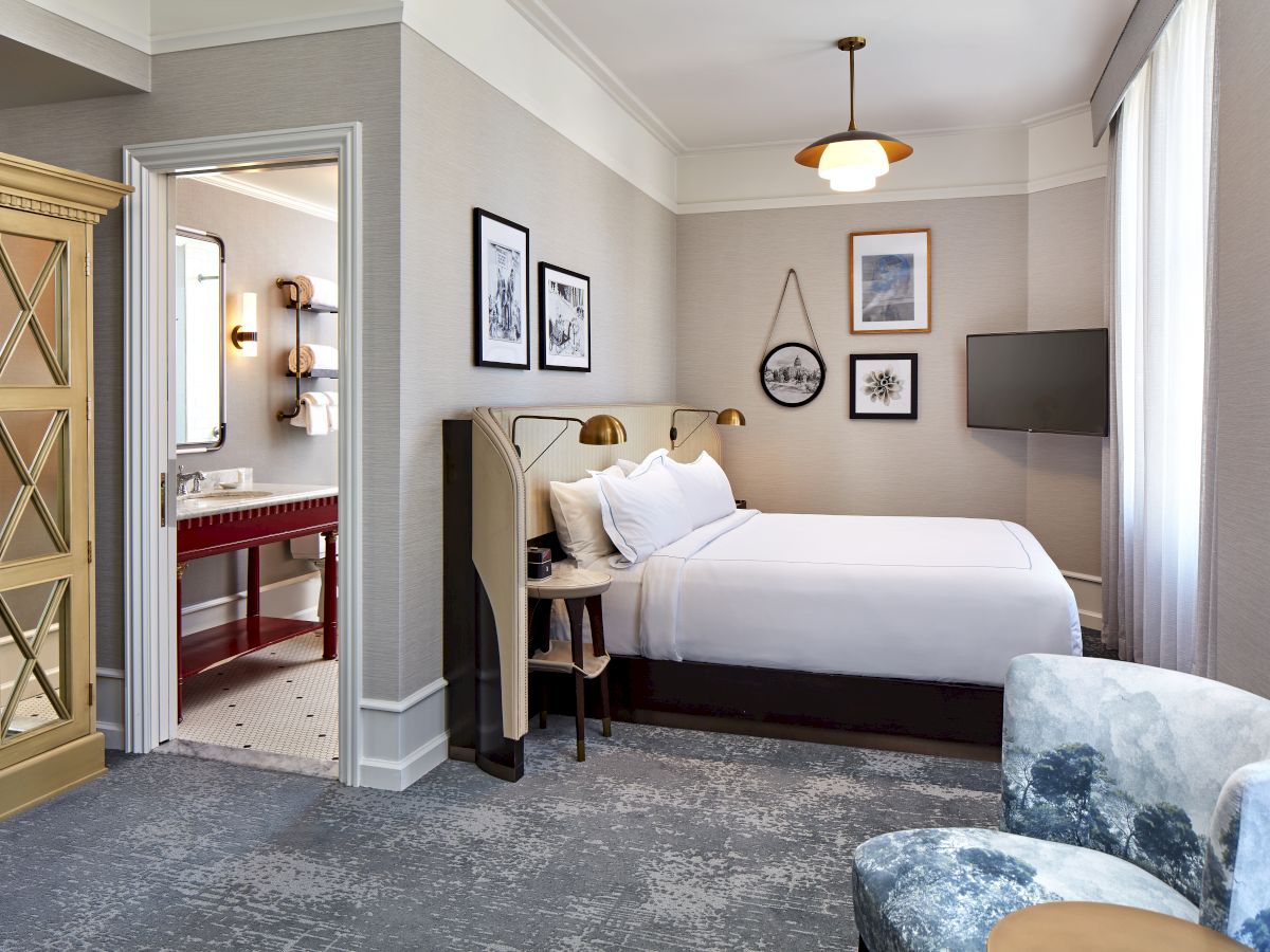 A modern hotel bedroom with a double bed, TV, framed art, pendant lights, and an open door leading to a bathroom.