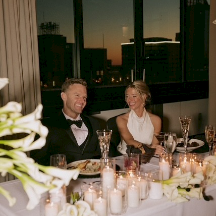 A dressed-up couple sits at a candlelit banquet table indoors, smiling with drinks in front of a nighttime city skyline.