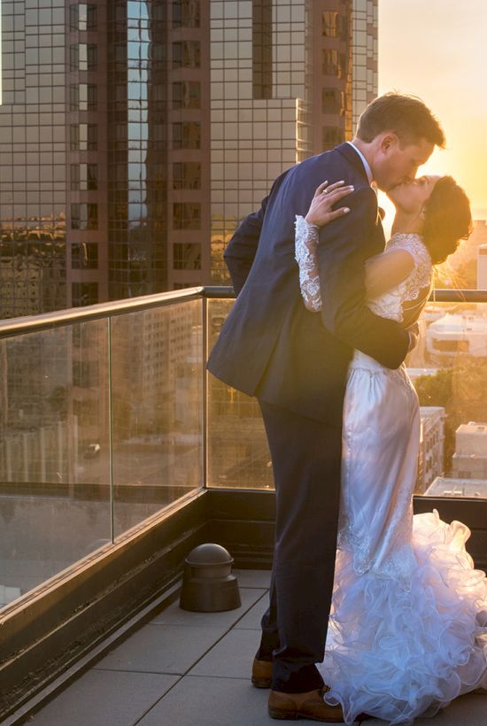 A groom and bride embrace on a rooftop balcony at sunset, framed by city skyscrapers and golden light.