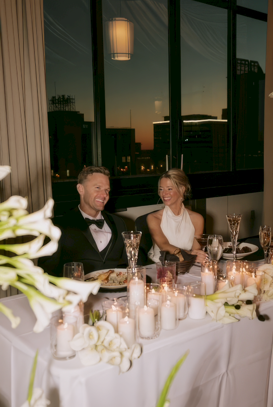 A couple in formalwear sits at a candlelit banquet table with glasses and flowers, gazing out at the city at dusk.