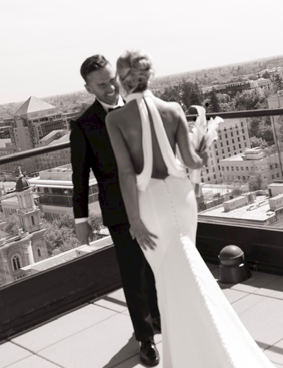 A black-and-white photo of a couple in wedding attire posing on a rooftop terrace with a city skyline behind them.
