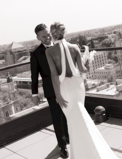 A smiling groom in a suit and his bride in a white dress laugh on a rooftop with a city view behind them.
