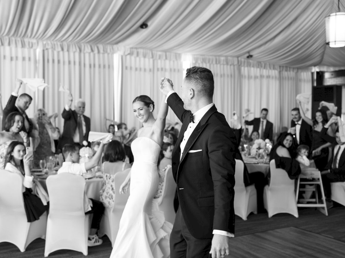 A couple dances at a wedding reception, with guests cheering under draped tent lights and hanging chandeliers.