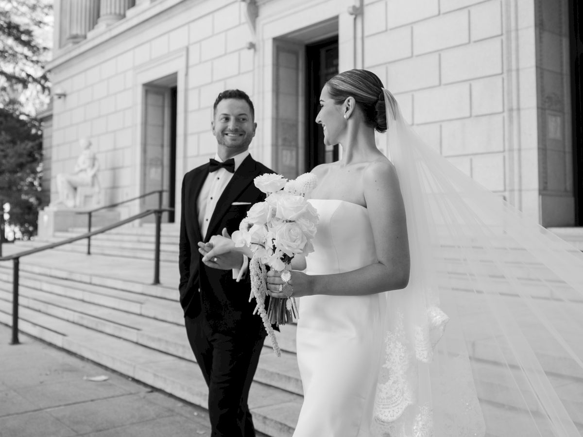 A bride and groom in formalwear pose outdoors, smiling with the bride&rsquo;s bouquet in front of a grand building.