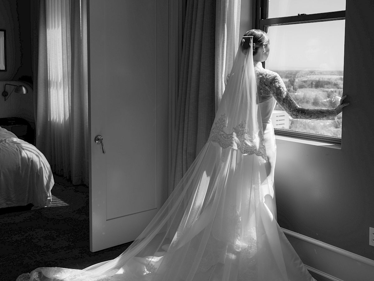 A woman in a white wedding gown and veil stands by a window, holding a curtain as sunlight pours in.