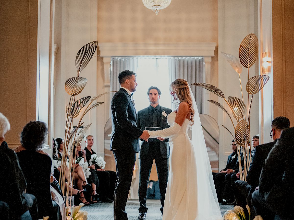 Wedding ceremony inside a hall: a couple holds hands and says vows on a flower-strewn aisle as guests watch.