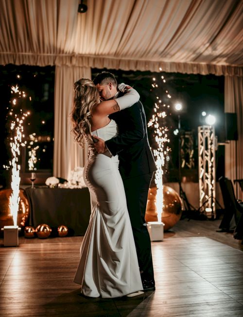 A bride and groom kiss under a tented wedding arch as sparklers burst around them, creating a sparkling celebration atmosphere.