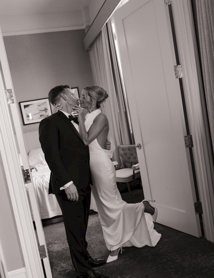 A black-and-white wedding photo shows a couple kissing in a doorway, the bride in a white gown and the groom in a suit, smiling.