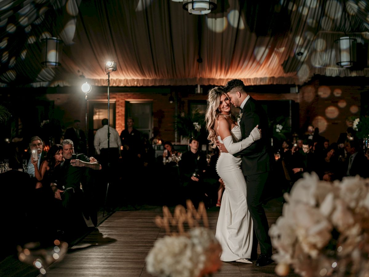 A newlyweds couple in formalwear slow-dancing at their reception while guests watch and phones light up the room.