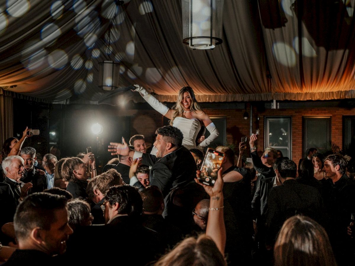 At a wedding reception, a woman in a white dress is lifted on shoulders as guests cheer and take videos.