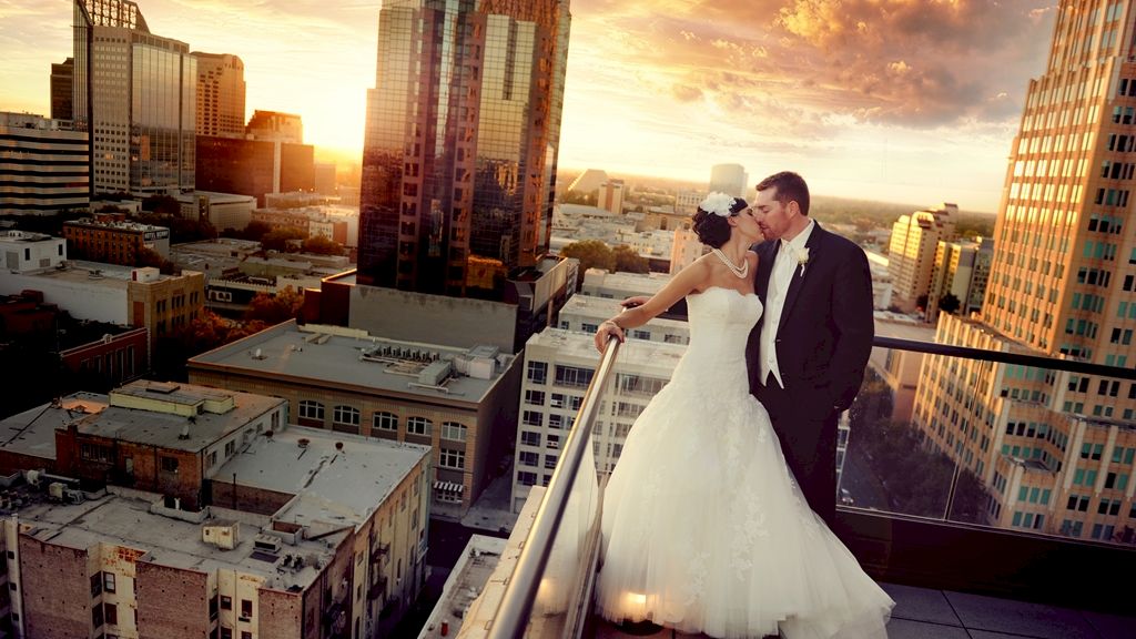 A bride and groom kiss on a rooftop at sunset above a city skyline, dressed for their wedding.