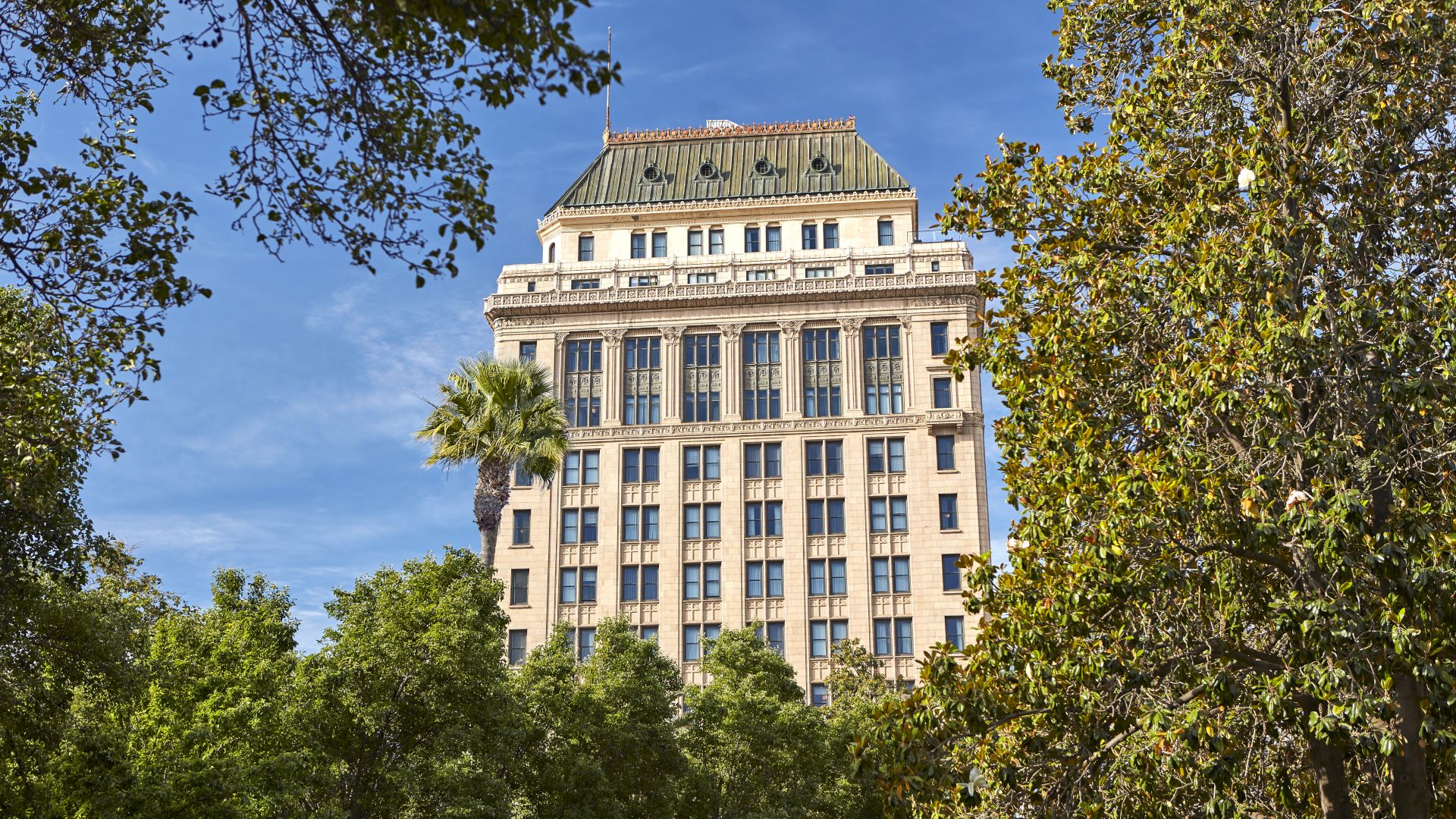 A tall historic office building rises behind leafy green trees under a bright blue sky.