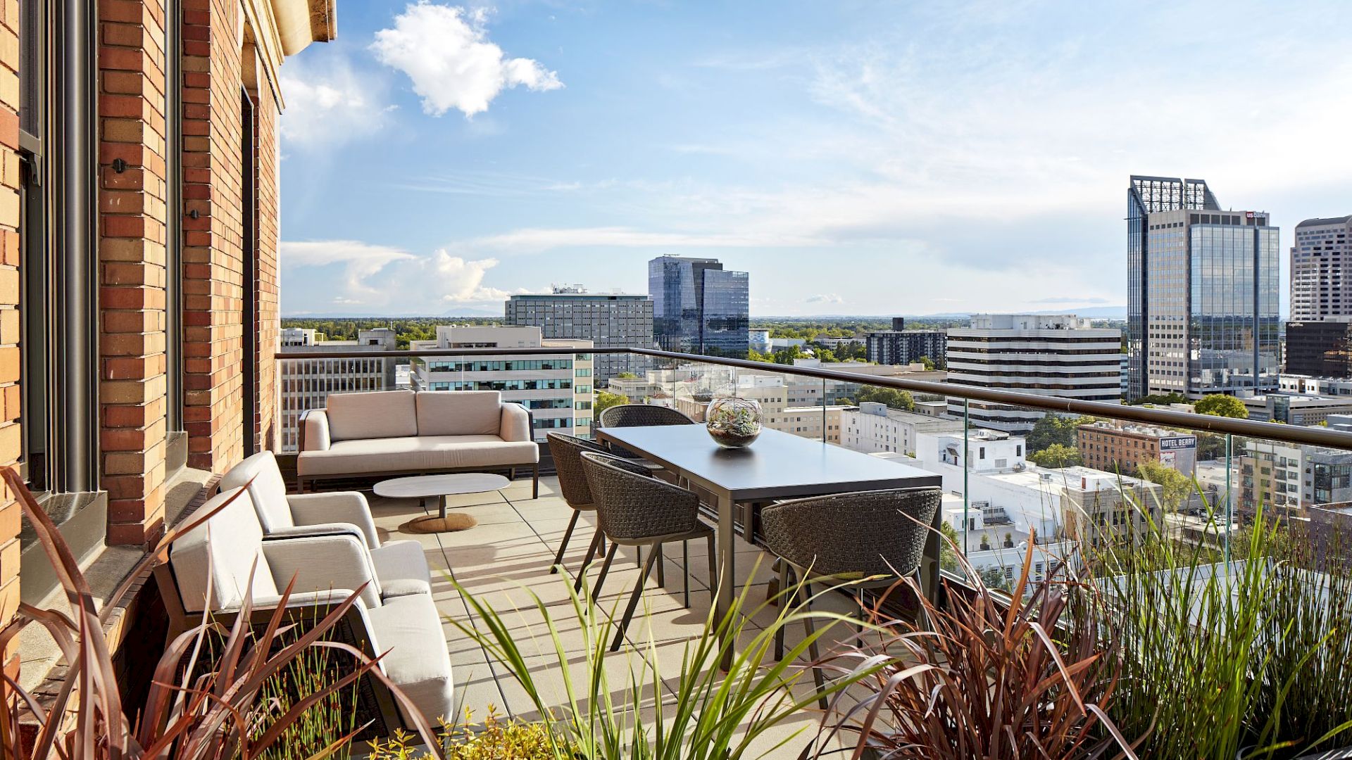 A sunny high-rise balcony with comfy outdoor seating and table, overlooking a city skyline and office buildings.