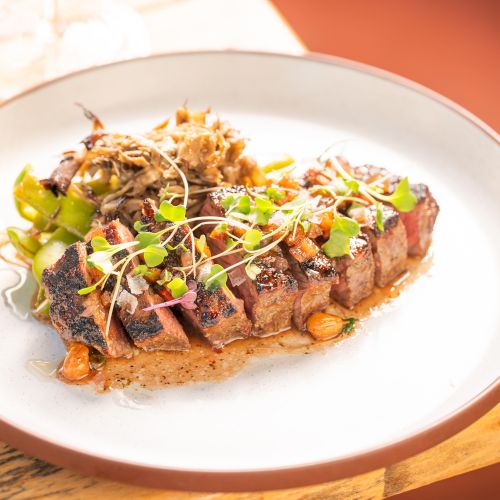 A plated steak with roasted vegetables, drizzled sauce, and bright microgreens on top, served on a white dish.