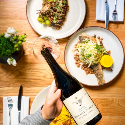 Two plated savory dishes with greens, plus a hand holding a wine bottle beside forks and glasses at a dining table.