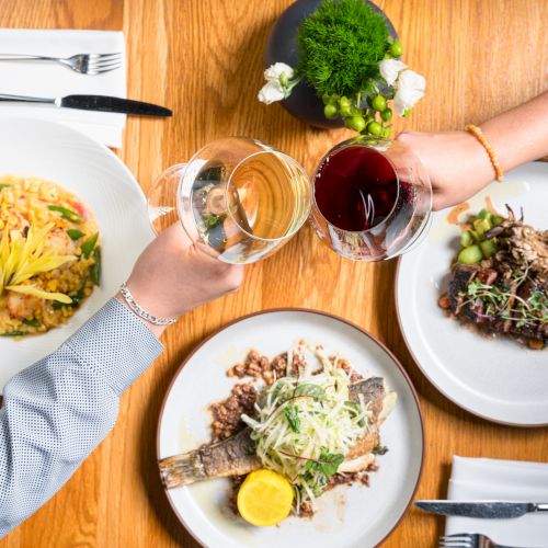 Friends toast over dinner, clinking wine glasses above plated entrees and sides at a table with cutlery nearby.