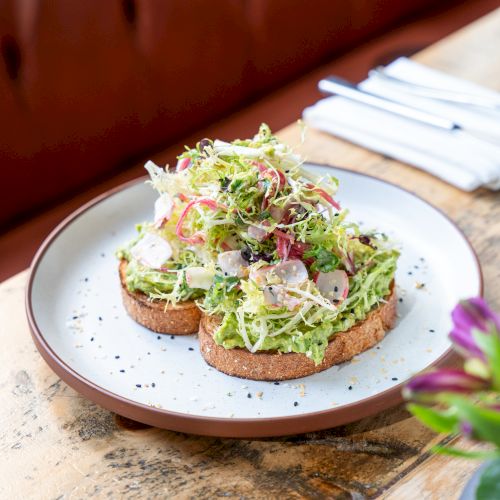 Two slices of toast topped with avocado, mixed greens, radish, and herbs, served on a plate with a fork nearby.