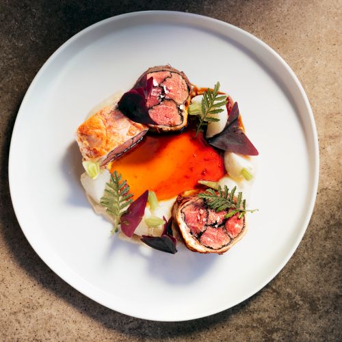 A white plate holding rolled beef slices with grilled mushrooms, purple herbs, and creamy root veggies in a red-orange sauce.