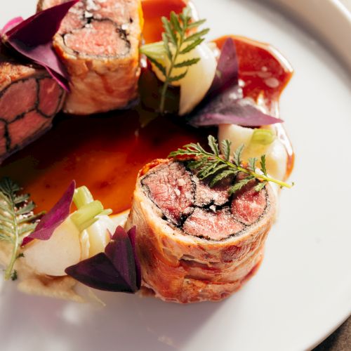 A plate of beef roulade in rich tomato sauce, topped with purple onion petals and fresh herbs beside creamy vegetables in gravy.