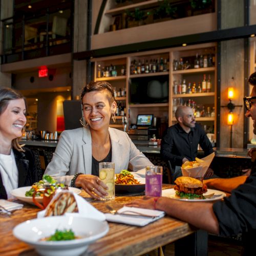 Three people dine and laugh at a restaurant table, with drinks and food in front of a lit bar.