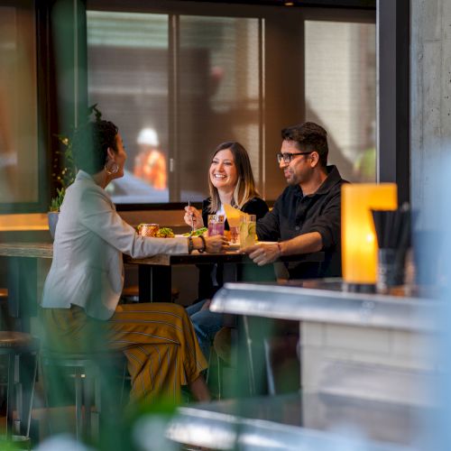 A group of friends chats and drinks at a restaurant table, smiling as a waiter serves food.
