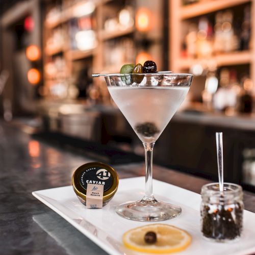 A chilled martini with two olives sits on a bar tray, with a caviar tin and blurred lounge lights behind it.
