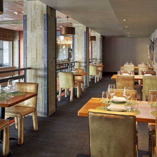 A modern restaurant dining room with tables set for dinner, pendant lights, bare concrete beams, and wall art.