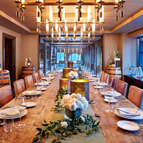 An elegant restaurant banquet table set with wine glasses, flowers, and barrels, lit by hanging copper chandeliers.